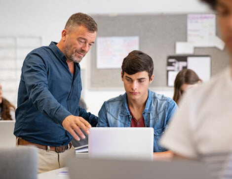 Teacher Shortage | Career Calling International A teacher assists a student with work on a laptop in class, highlighting the need for quality vet compliance.