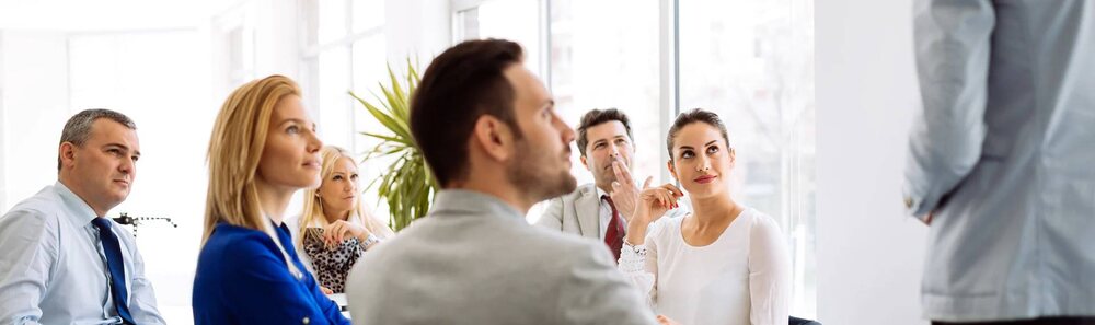 Business professionals engage with a presenter during an office meeting, reflecting our education consultancy services.